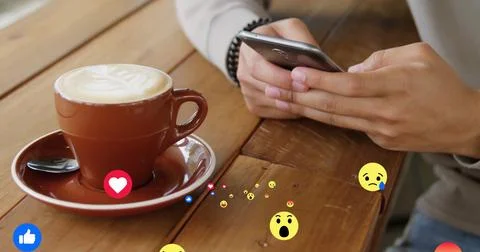 Holding smartphone tapping screen at coffee shop table, with latte cup and emoji 写真素材