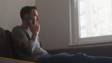 Home office worker sit on couch with laptop on his lap and take call on phone Stock Photos