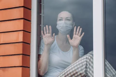 Home quarantine. A Caucasian woman is sitting at window in a medical mask Stock Photos