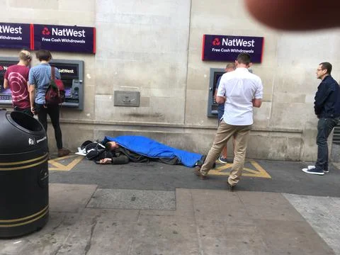 Homeless person sleeping rough while people withdraw cash from ATM machines. Stock Photos