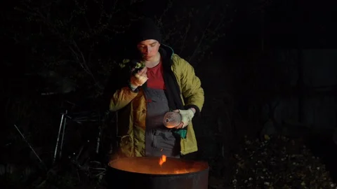 A homeless young man stands by the fire and eats an apple. A man stands at night Stockbeeldmateriaal 129557891