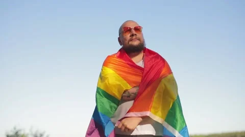 Homosexual bold man wrapped in a rainbow gay flag while parade on background of Видео 134813288