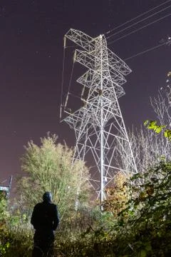 A hooded figure, back to camera. Looking up at a electricity pylon Stock Photos