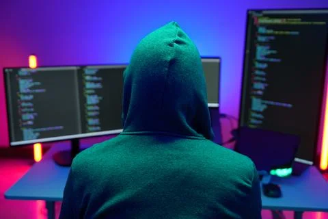 Hooded hacker coding at dual monitors in neon lit room Stock Photos