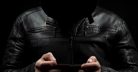 A hooded hacker with a leather jacket holding a smart phone with his both hands Video stock 328319776