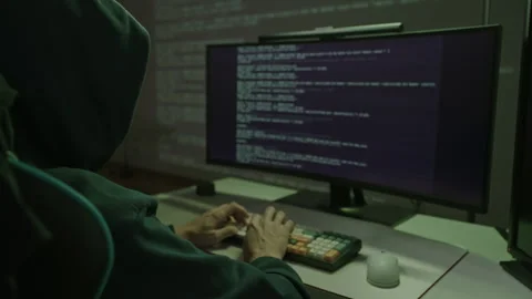 Hooded hacker person typing on computer keyboard in dark room, engaging in cyber Stock Footage 323449480