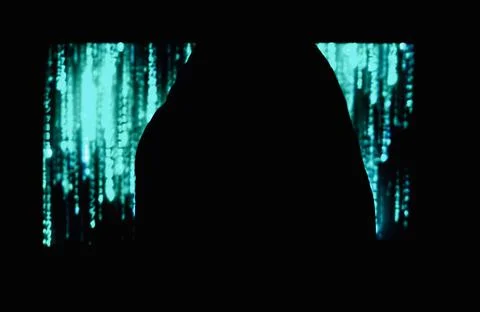 Hooded hacker sitting in front of a computer screen with green code. 스톡 사진