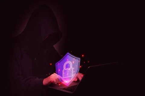 A hooded hacker types on a laptop, with a security shield representing prot.. Foto stock