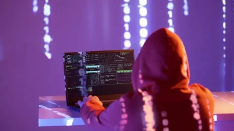 A hooded hacker types on a laptop surrounded by projected binary code and 動画素材 307008063