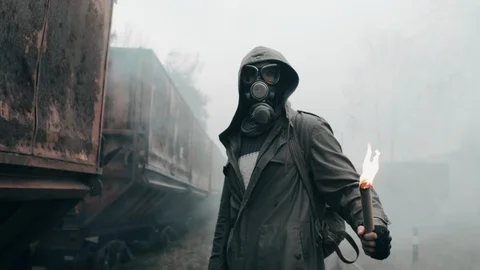 Hooded man in gas mask standing by abandoned carriages with burning fire in hand Stock Footage