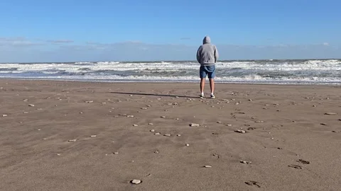 Hooded man standing alone on beach looking at ocean waves. 4K Stock Footage 329680995