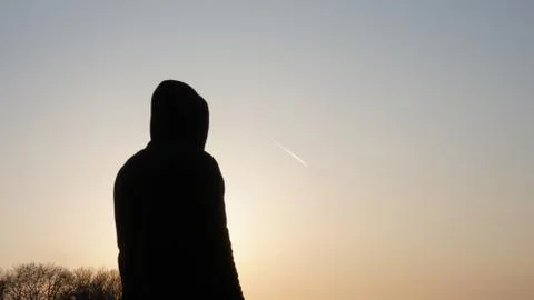A hooded man standing, back to camera. Watching a plane with a contrail Foto stock