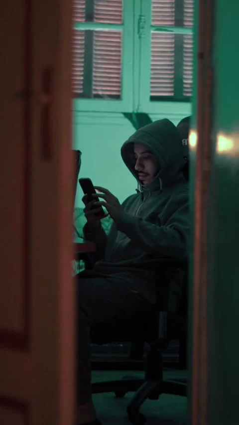 Hooded Man Using Phone in Dimly Lit Room Viewed Through Door Stock Footage 309770753