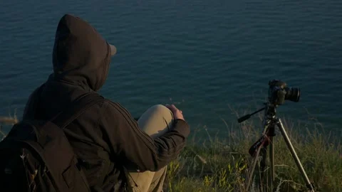 Hooded person with a camera sitting in front of the sea in slowmotion Stock Footage 188506835