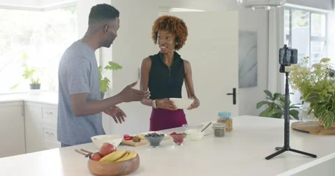 Hosts grey tee, black crop top demoing at kitchen island holding bowl, phone on Stock Footage 339415161