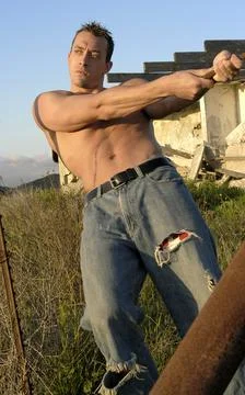 Hot athletic young man poses shirtless pulling a cable on a broken down farm. Stock Photos