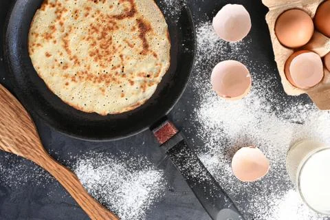Hot pancake in black pan on black table with flour, milk and eggs Stock Photos