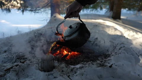 Hot tea from a camping kettle is poured into a metal mug standing on the snow Stock Footage 329968835