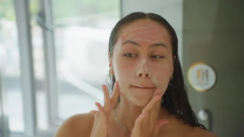 Hotel bathroom guest gently exfoliating face at mirror with fine scrub, slow Stock Footage 330042453