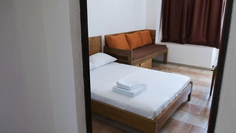 A hotel room with a white double bed, brown sofa with orange cushions and 스톡 동영상 89914366