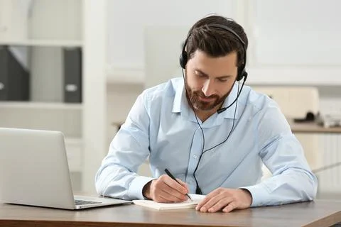Hotline operator with headset writing something in notebook while working a.. Stock Photos