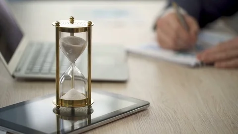 Hourglass standing on table with sand trickling, man working with graphs, data Stock Footage