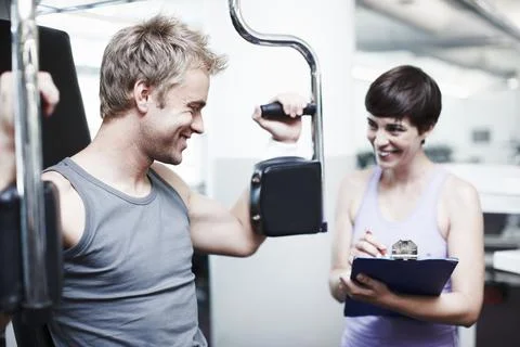 How am I doing, coach. a handsome young man using an exercise machine while his Stock Photos