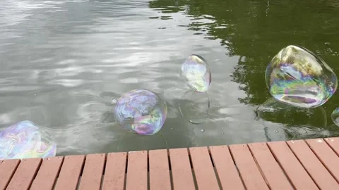 Huge rainbow soap bubbles float in air along a red lakeside dock Video stock 233740270