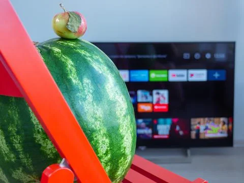 Huge watermelon and tiny apple watching TV set. Stock Photos