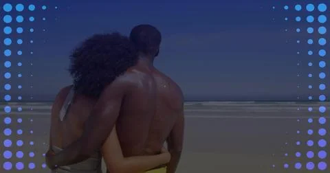 Hugging pair standing on sandy shore facing ocean, showing swimsuit strap, Fotos Stock