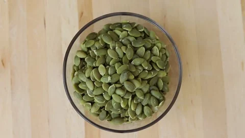 Hulled green pumpkin seeds falling into a clear glass bowl Stock Footage 346444613