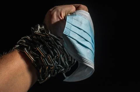 Human hand chained in chains holds a medical protective mask, isolated on black  Stock Photos