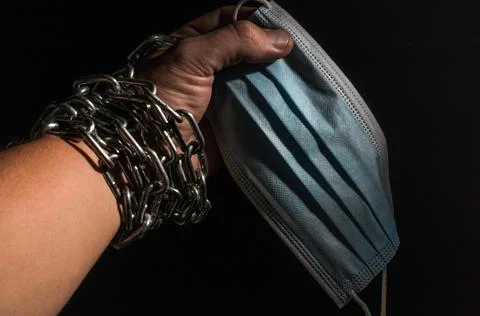 Human hand chained in chains holds a medical protective mask, isolated on black  Stock Photos