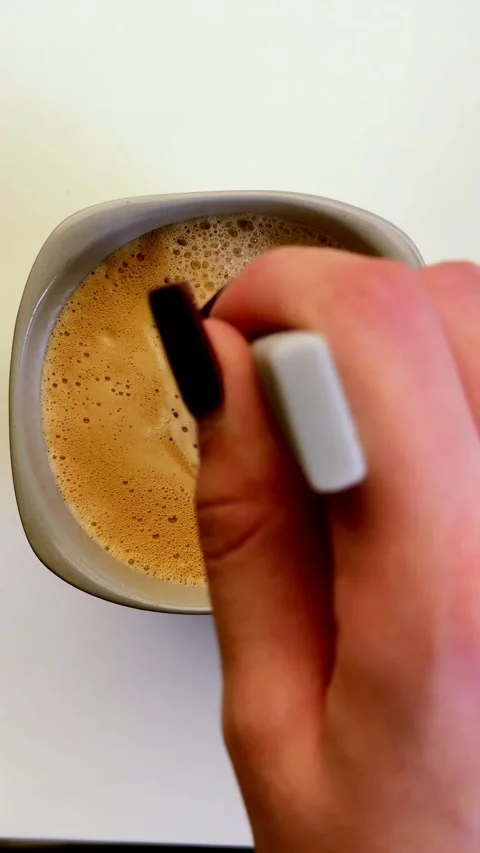 Human hand gently stirs a frothy coffee drink, blending rich brown liquid with Stock Footage 335951915