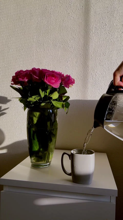 Human hand pours liquid from clear kettle into white cup on table near blooming Video stock 333631569