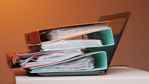 Human hands stack a towering pile of colorful binders and paperwork onto an Stock Footage 347237106