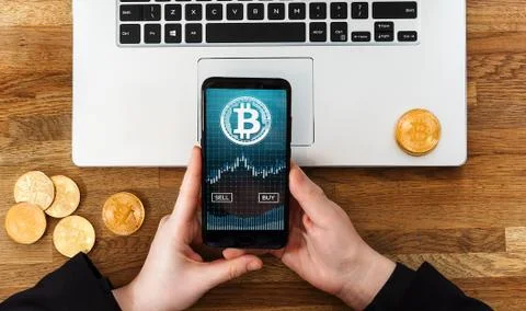 Human using smartphone app to buy or sell bitcoins. Laptop and gold coins of Stock Photos