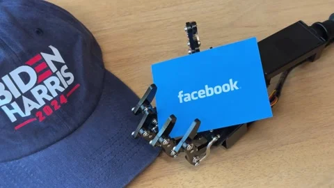 Humanoid Robot Hand Holding Facebook Card Near Biden Harris Hat Stock Footage 280009678