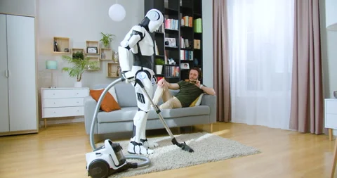 Humanoid robot vacuuming carpet while man in casual wear lying on grey sofa at Stock Footage 244137638