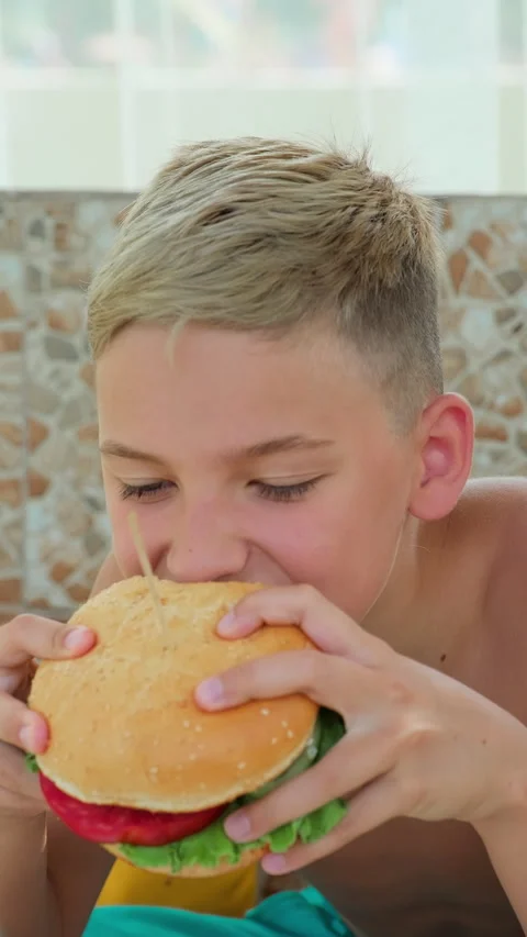 Hungry blond boy eating hamburger with lettuce and tomato in a sunny outdoor Stock Footage 313740824