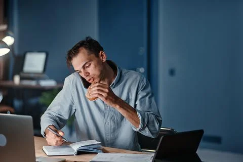 Hungry businessman snacking on a hamburger while working at night. Stock Photos
