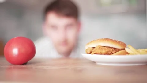 Hungry man chooses between a cheeseburger and a tomato. Fast food or vegetable Stock Footage 132355574