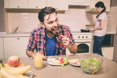 Hungry man is sitting at table and biting meal he has on fork. Guy is keeping Stock Photos