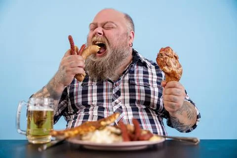 Hungry plus size man eats sausages at table with rich food and mug of beer Foto stock