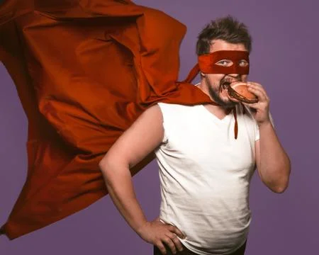 Hungry superhero man eats big hamburger with meat. Man in red flying cloak eats Stock Photos