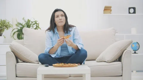Hungry woman is switching on the TV program, while sitting on a sofa. She is Vídeos de archivo 120884678