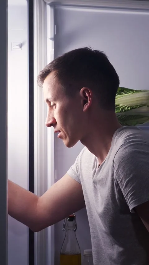 Hungry young man opening refrigerator at night, eating a sweet pastry and Stock Footage 342904378