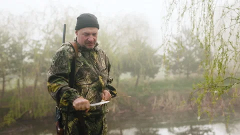 A hunter with a sharp knife sharpens a stick near a misty autumn lake Stock Footage 196623161