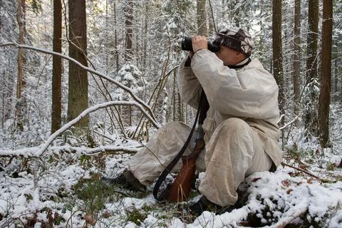 A hunter sits on a fallen tree and looks through binoculars in the winter for Stock Photos