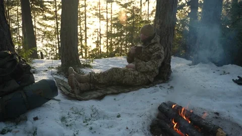 The hunter sits by the fire in the winter forest and drinks from a mug Видео 234321819